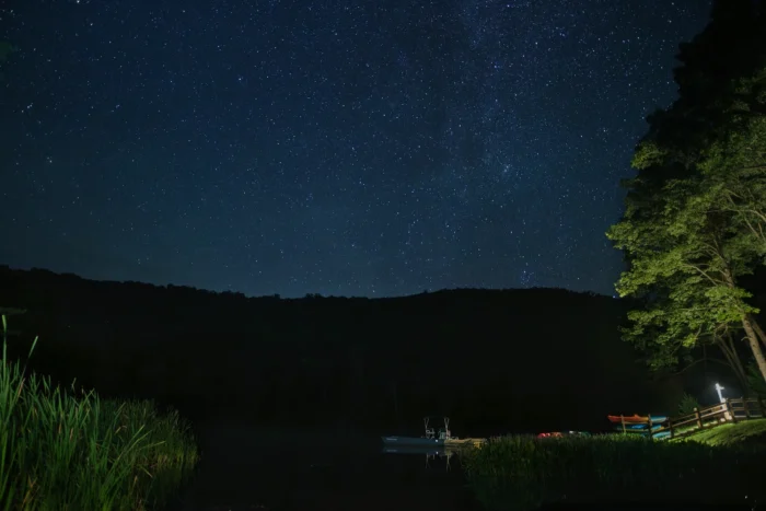Star filled night sky over a dark mountain ridge at Watoga State Park, with a small dock, boats and glowing lamp post along a quiet lake shore.