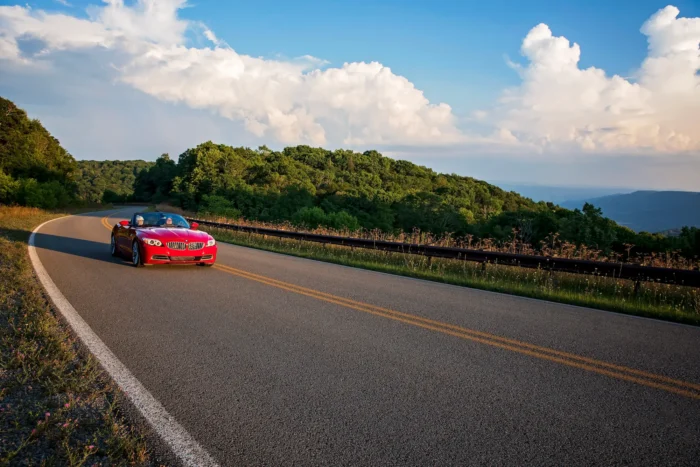 Red convertible driving on the curving Highland Scenic Highway lined with trees, overlooking distant hills under a partly cloudy sky.