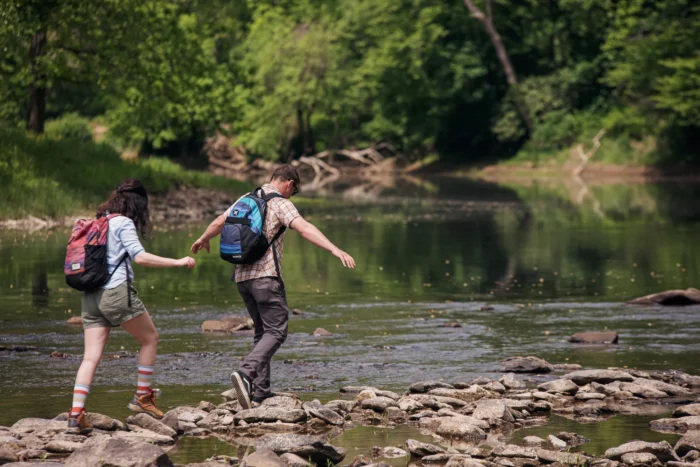 Two hikers with backpacks balancing on rocks while crossing the calm Greenbrier River.