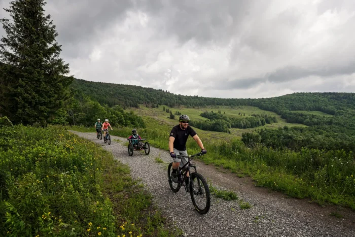 Group of cyclists riding gravel bikes down a hilly trail through green meadows of the Mower Basin in the Monongahela National Forest, with a man smiling in front and rolling forested hills behind.