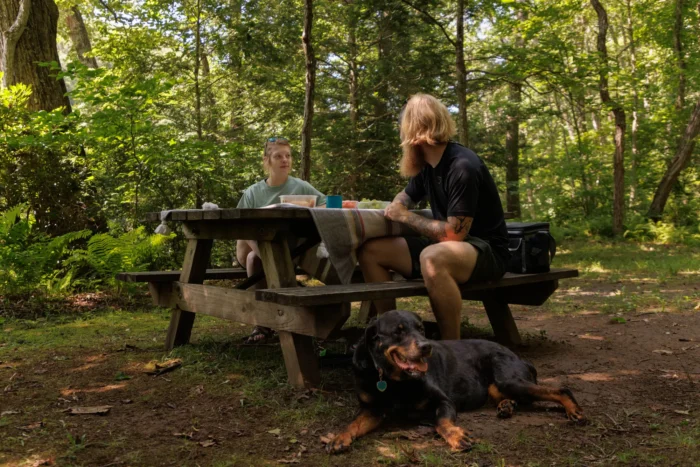 Two people sitting at a picnic table in the woods with their dog.