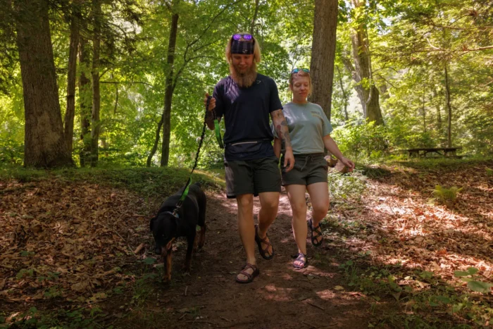A couple hikes along a forested trail with their leashed dog. 