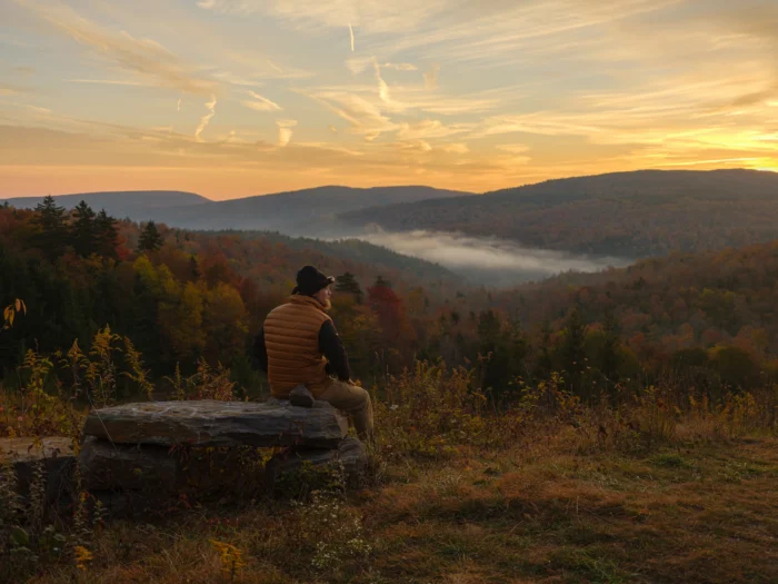 A man sitting on a large rock watches the sunrise over the mountains covered in fall foliage. 