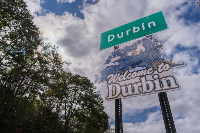 A Welcome to Durbin sign stands beside trees with clouds overhead. 