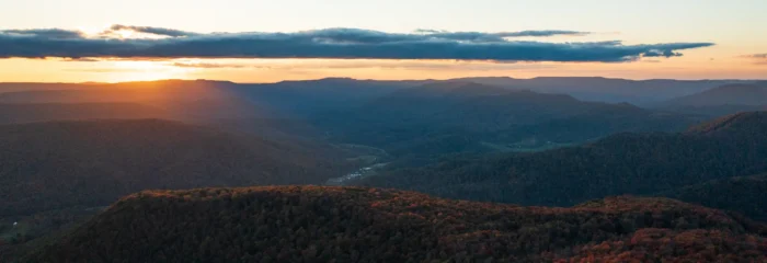 Panoramic sunset view over rolling forested mountains with warm golden light and dramatic clouds on the horizon.