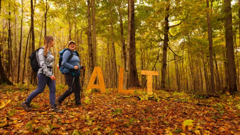 Two women hike past the ALT sign in the forest, with fallen leaves on the ground.