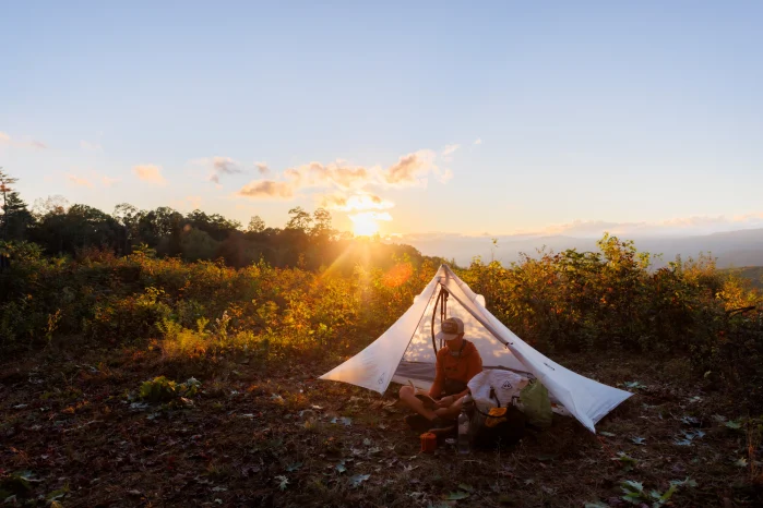 A hiker sits under a white tent along the Allegheny Trail. The sun is setting behind him. 